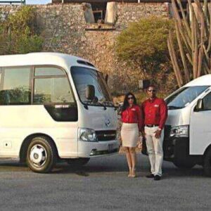 Two people in red shirts and light bottoms stand between a white minibus and van, awaiting their Shared Airport Transfer Zone C amid sunny stone walls, desert plants, and a partially visible building above the wall.