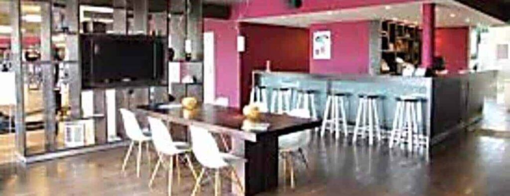 Modern interior with a wooden table, four white chairs, and a bar with high stools. Watch games on the wall-mounted TV and enjoy your Short Term sport Membership. Features dark wood flooring, red accent walls, and decorative shelves.