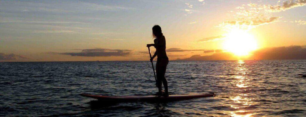 During the Spanish Water Tour, a person stands on a paddleboard in calm ocean waters at sunset, with warm hues in the sky, gentle waves reflecting sunlight, and distant mountains on the horizon.