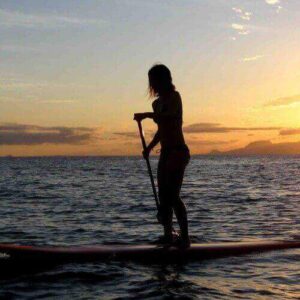 During the Spanish Water Tour, a person stands on a paddleboard in calm ocean waters at sunset, with warm hues in the sky, gentle waves reflecting sunlight, and distant mountains on the horizon.