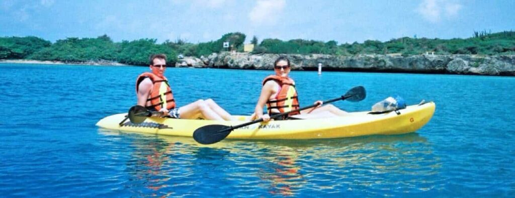 Twee mensen in oranje reddingsvesten peddelen in een gele tandemkajak op helderblauw water tijdens de "Kajaktocht bij zonsondergang - Spaans Water," met weelderig groen en een heldere wolkenlucht op de achtergrond.