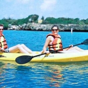 Two people in orange life jackets paddle a yellow tandem kayak on the bright blue Spanish Water, sunglasses on, gliding past a green, rocky shore under a partly cloudy sky—perfect for the Sunset Kayak Tour - Spanish Water -.