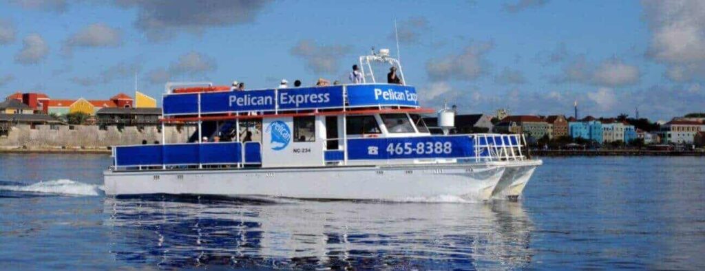 The Sunset the West, a blue and white passenger boat, cruises west on calm water near a pastel-colored coastal town at sunset, with its phone number 465-8388 displayed on the side.