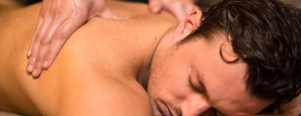 Een man met kort donker haar ligt met zijn gezicht naar beneden, zijn ogen gesloten, terwijl hij geniet van een kalmerende Zweedse massage. De masseuse drukt zachtjes op zijn bovenrug, waardoor een kalmerende en ontspannen sfeer ontstaat.