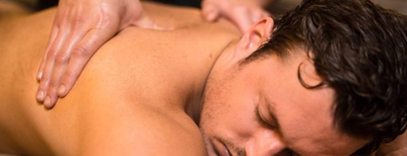 A man with short dark hair lies face down, eyes closed, as two hands gently massage his oiled upper back during a Swedish Relax Massage. The atmosphere is calm and soothing.