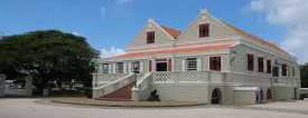 The Curaçao Museum is in a two-story colonial-style building with light gray walls, red roof tiles, and red shutters. A large staircase leads to its porch, with trees and a mostly clear blue sky in the background.