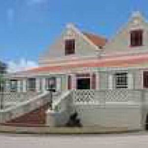The Curaçao Museum is in a two-story colonial-style building with light gray walls, red roof tiles, and red shutters. A large staircase leads to its porch, with trees and a mostly clear blue sky in the background.