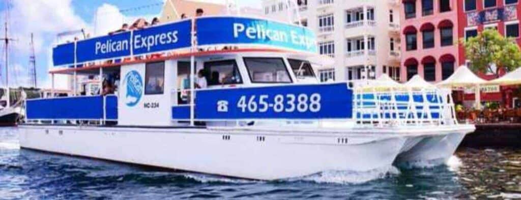 De dubbeldekkerboot "Pelican Express", wit en blauw met opvallende borden en 465-8388, vaart op een heldere, zonnige dag door De gelukkige haven langs kleurrijke gebouwen aan het water en zitjes in de openlucht.