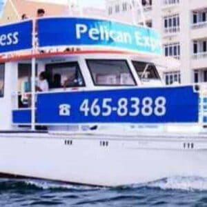A white and blue catamaran named Pelican Express cruises past The Happy Harbour, a popular seafood spot by colorful waterfront buildings, displaying the phone number 465-8388 under a clear sky.