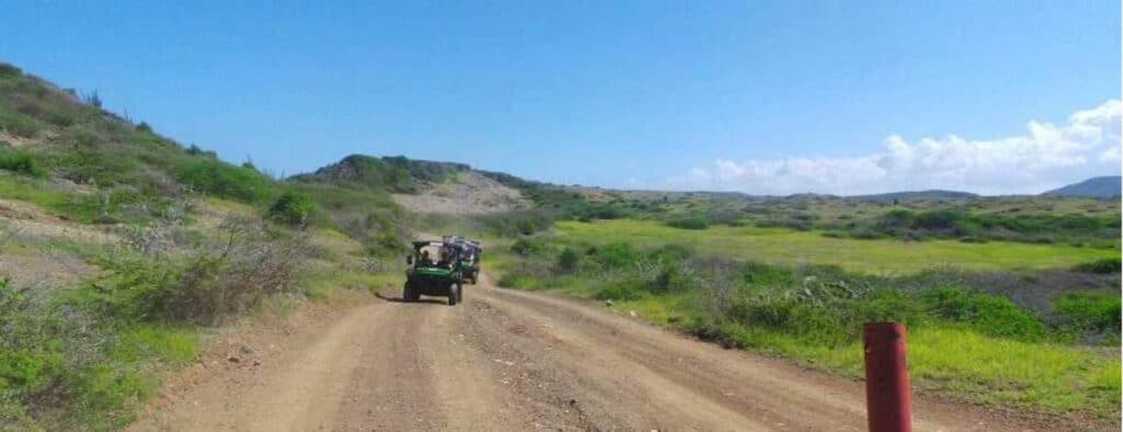 A Lazy Beach Off-Road tour vehicle travels along a dirt road through green hills beneath a clear blue sky, with passengers inside. Low shrubs and grass border the route, and a red post is seen in the foreground.