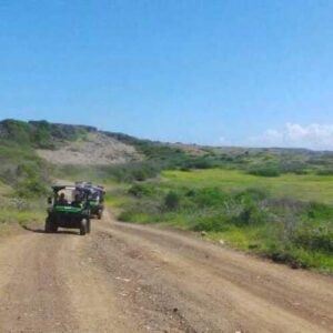 A Lazy Beach Off-Road tour vehicle travels along a dirt road through green hills beneath a clear blue sky, with passengers inside. Low shrubs and grass border the route, and a red post is seen in the foreground.
