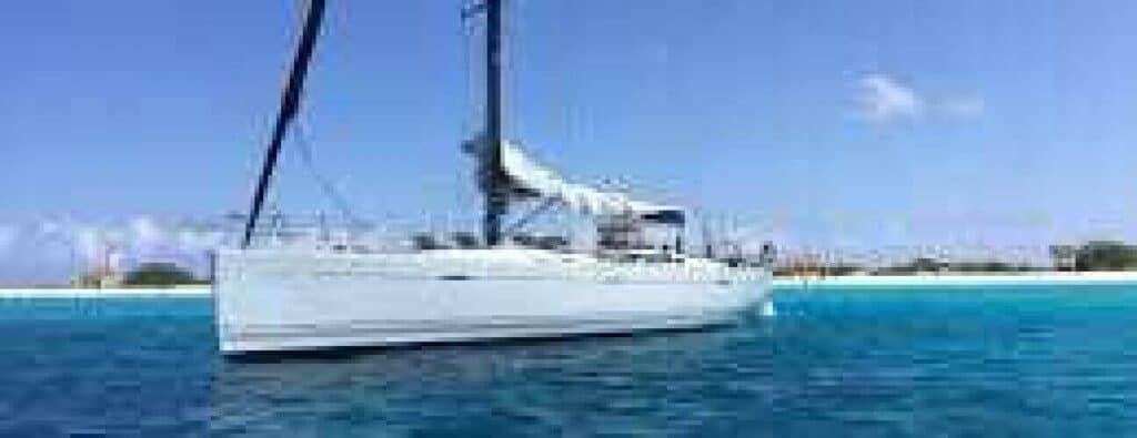 A white sailboat with a tall black mast floats on clear blue water near a sandy beach, making the TransMark Sunset tour ideal. The shoreline and greenery appear in the background beneath a bright blue sky.
