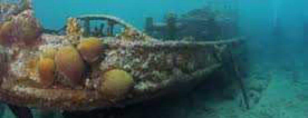 A submerged, rusted car encrusted with barnacles and marine growth rests near the TRIP TUGBOAT & FUIK wreck, surrounded by blue-green water and natural sea life in low visibility on the ocean floor.