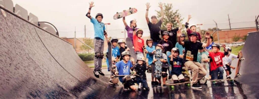 A lively group of kids and teens in helmets and pads pose with skateboards on a wet ramp at Vacation Camps. Some are mid-jump and cheering, joined by a few adults, with trees and a brick building in the background.