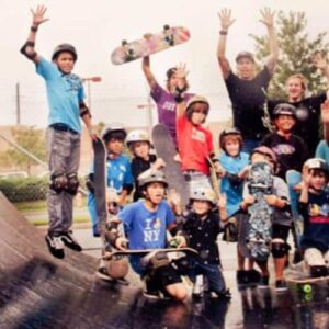 A lively group of kids and teens in helmets and pads pose with skateboards on a wet ramp at Vacation Camps. Some are mid-jump and cheering, joined by a few adults, with trees and a brick building in the background.