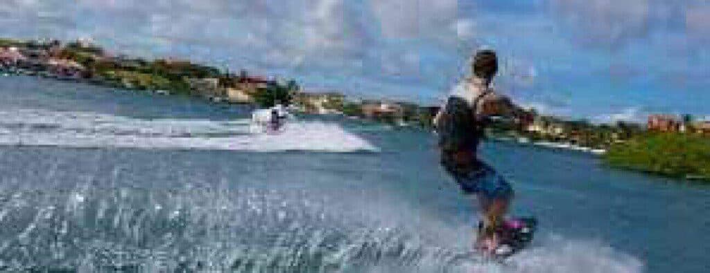 Iemand in een zwemvest en korte broek berijdt de Wakeboarden Vissen en duiken op een zonnige dag, terwijl hij een touw vasthoudt achter een speedboot met opspattend water, stadsgebouwen, bomen en een gedeeltelijk bewolkte lucht op de achtergrond.