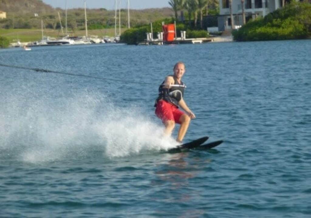 A man enjoying waterskiing wears a black life vest and red shorts, smiling as he glides on a lake while being pulled by a rope. Water splashes behind him, with boats, docks, and waterfront buildings amid greenery in the background.