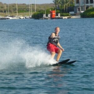 A man enjoying waterskiing wears a black life vest and red shorts, smiling as he glides on a lake while being pulled by a rope. Water splashes behind him, with boats, docks, and waterfront buildings amid greenery in the background.