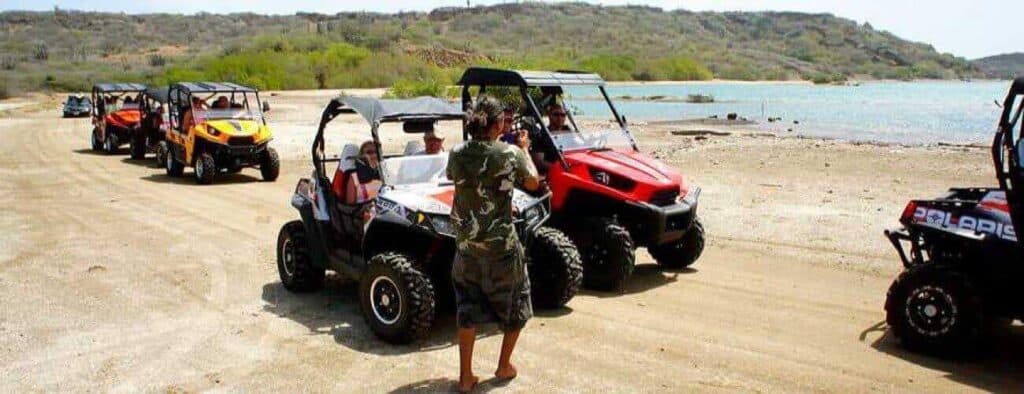 Several West Tour by buggy vehicles, each with passengers, are parked in a row on a sandy path near water. A person in shorts and a camo shirt stands at the front, likely giving instructions as green hills rise under the bright sky.