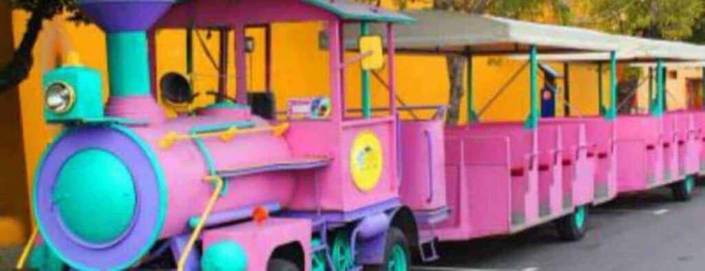The Willemstad Tour by Trolley Train features a brightly colored pink and turquoise train with open passenger cars, parked on a paved road beside a yellow wall, creating a cheerful, playful vibe for your sightseeing adventure.