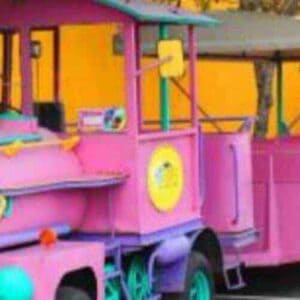 De Rondleiding door Willemstad met de trolleytrein heeft een groenblauwe en roze locomotief met openluchtauto's voor kinderen, geparkeerd in een straat bij een levendige gele muur en groene bomen, die een vrolijke, speelse sfeer creëren.