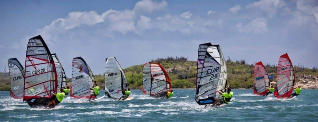A group of windsurfers in neon green shirts, likely taking Windsurfing Lessons, race across choppy blue water with numbered sails against a sandy shore lined with green bushes and a partly cloudy sky.