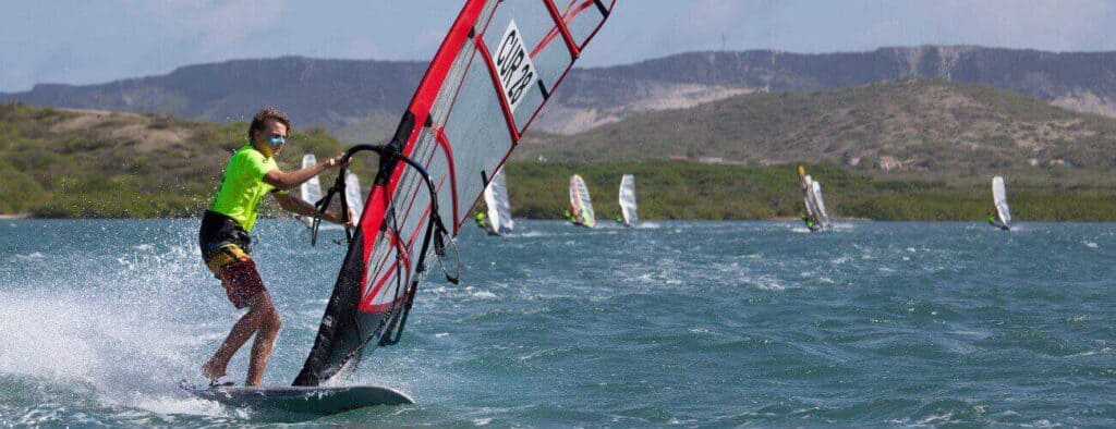A windsurfer in a bright green shirt glides swiftly on blue water, creating a spray. Others enjoy Windsurfing Rental nearby under a sunny sky, with green hills and mountains in the distance.