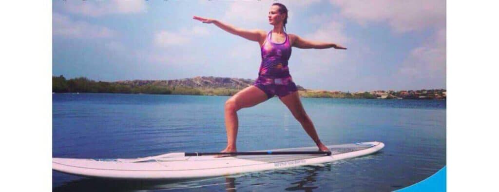 Wearing a purple Yunayoga outfit, a woman practices Warrior II on a paddleboard floating on calm water, arms extended as a paddle rests across the board. Blue sky, distant rocky hills, and greenery form the backdrop.