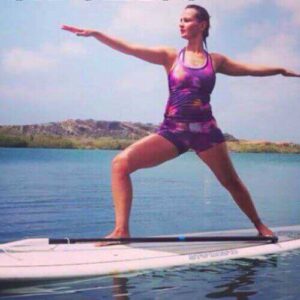 Wearing a purple Yunayoga outfit, a woman practices Warrior II on a paddleboard floating on calm water, arms extended as a paddle rests across the board. Blue sky, distant rocky hills, and greenery form the backdrop.