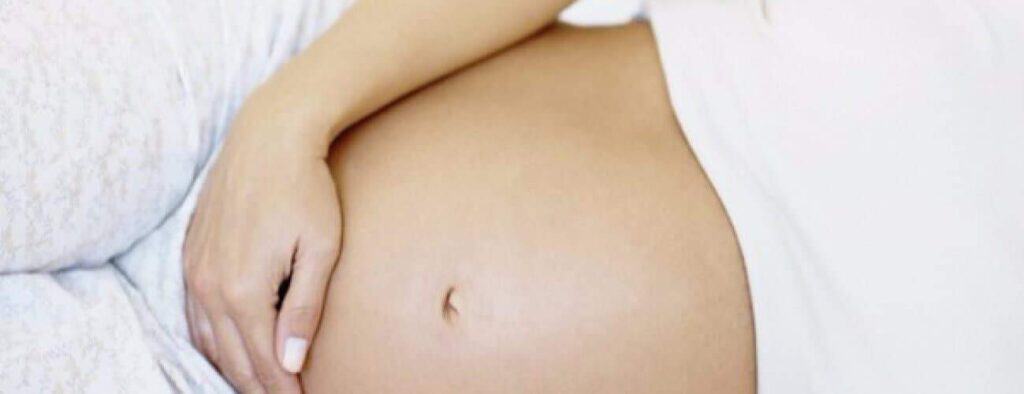 A relaxing Pregnancy Massage is shown with a close-up of a bare baby bump, a hand gently resting on the side. The blurred background and light-colored clothing highlight comfort and calm for expecting mothers.