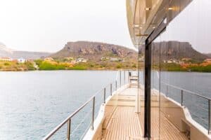Tijdens een Varen in pure luxe: Gold Seahorse privétour, glijdt het jacht met een houten dek en metalen reling over kalme wateren langs rotsachtige, groene heuvels omzoomd met huizen, waarbij de glazen wand het verbluffende uitzicht weerspiegelt.