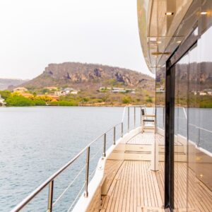 Tijdens een Varen in pure luxe: Gold Seahorse privétour, glijdt het jacht met een houten dek en metalen reling over kalme wateren langs rotsachtige, groene heuvels omzoomd met huizen, waarbij de glazen wand het verbluffende uitzicht weerspiegelt.