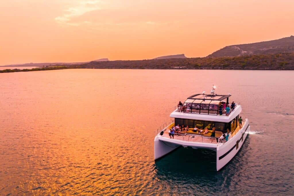 The Sunset Catamaran Yacht Trip in Curaçao features a large white catamaran sailing on calm water at sunset, with people relaxing on deck and vibrant orange-pink skies reflecting over tropical coastline and green hills.