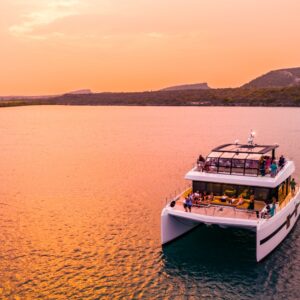 De Catamaran zeiltocht bij zonsondergang op Curaçao biedt een ontspannen tocht op een moderne witte catamaran met uitzicht op groene heuvels en de kustlijn, terwijl de lucht roze-oranje kleurt en passagiers genieten op het dek.