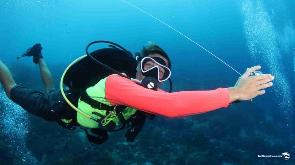 Wearing a neon green vest and red wetsuit, a scuba diver extends an arm underwater holding a device amid bubbles, with coral and blue ocean in the background—enjoy the 6-Day Unlimited Shore Diving Package.