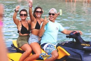 Three people smile and make peace signs while riding a yellow jet ski on the water. Two women in swimsuits and sunglasses sit behind a man in a blue shirt and cap. The Paranda Boat Trips in Curaçao logo is at the bottom center of the image.
