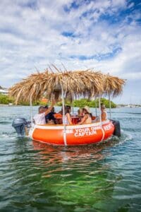 The Tiki BBQ Boat XL: Be Your Own Captain, a round orange boat with a thatched roof and outboard motor, carries six people on clear green water, surrounded by lush greenery and blue sky. Passengers chat and relax onboard.