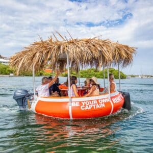 Un barco circular naranja Tiki BBQ Boat XL: Sé tu propio capitán con techo de paja flota sobre el agua clara, donde varias personas se relajan y charlan. La embarcación luce un cartel de alquiler, con un frondoso follaje verde y un cielo parcialmente nublado de fondo.