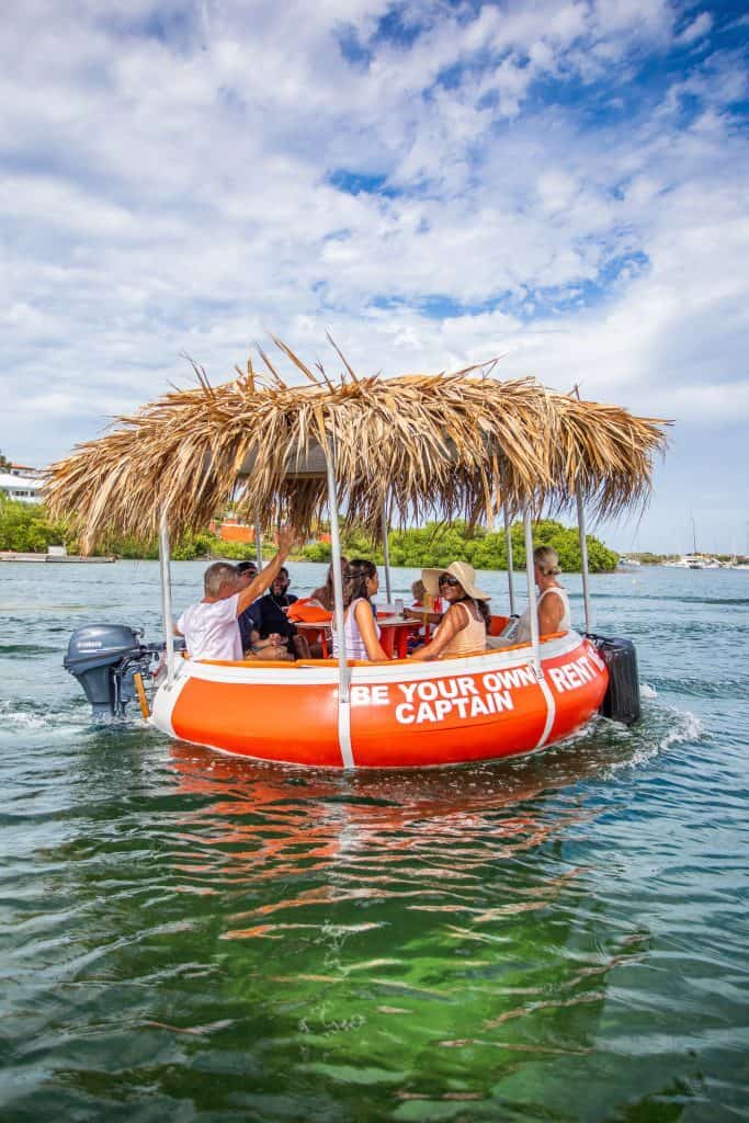 Un barco circular naranja Tiki BBQ Boat XL: Sé tu propio capitán con techo de paja flota sobre el agua clara, donde varias personas se relajan y charlan. La embarcación luce un cartel de alquiler, con un frondoso follaje verde y un cielo parcialmente nublado de fondo.