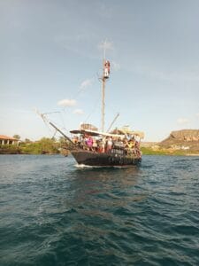 The Pirate Adventure tour boat “Simba,” styled like a pirate ship, cruises with passengers on blue-green water past green shores, houses, and distant rocky hills beneath a clear sky. Someone stands atop the mast.