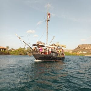 Piraten Avontuur is een zwarte rondvaartboot in piratenstijl genaamd "Simba", tropisch versierd, varend in blauwgroene wateren langs huizen, groene heuvels en een rotsachtige berg onder een heldere hemel met veel passagiers aan boord.