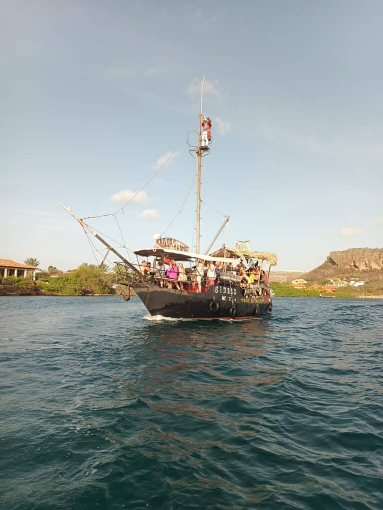 The Pirate Adventure tour boat “Simba,” styled like a pirate ship, cruises with passengers on blue-green water past green shores, houses, and distant rocky hills beneath a clear sky. Someone stands atop the mast.