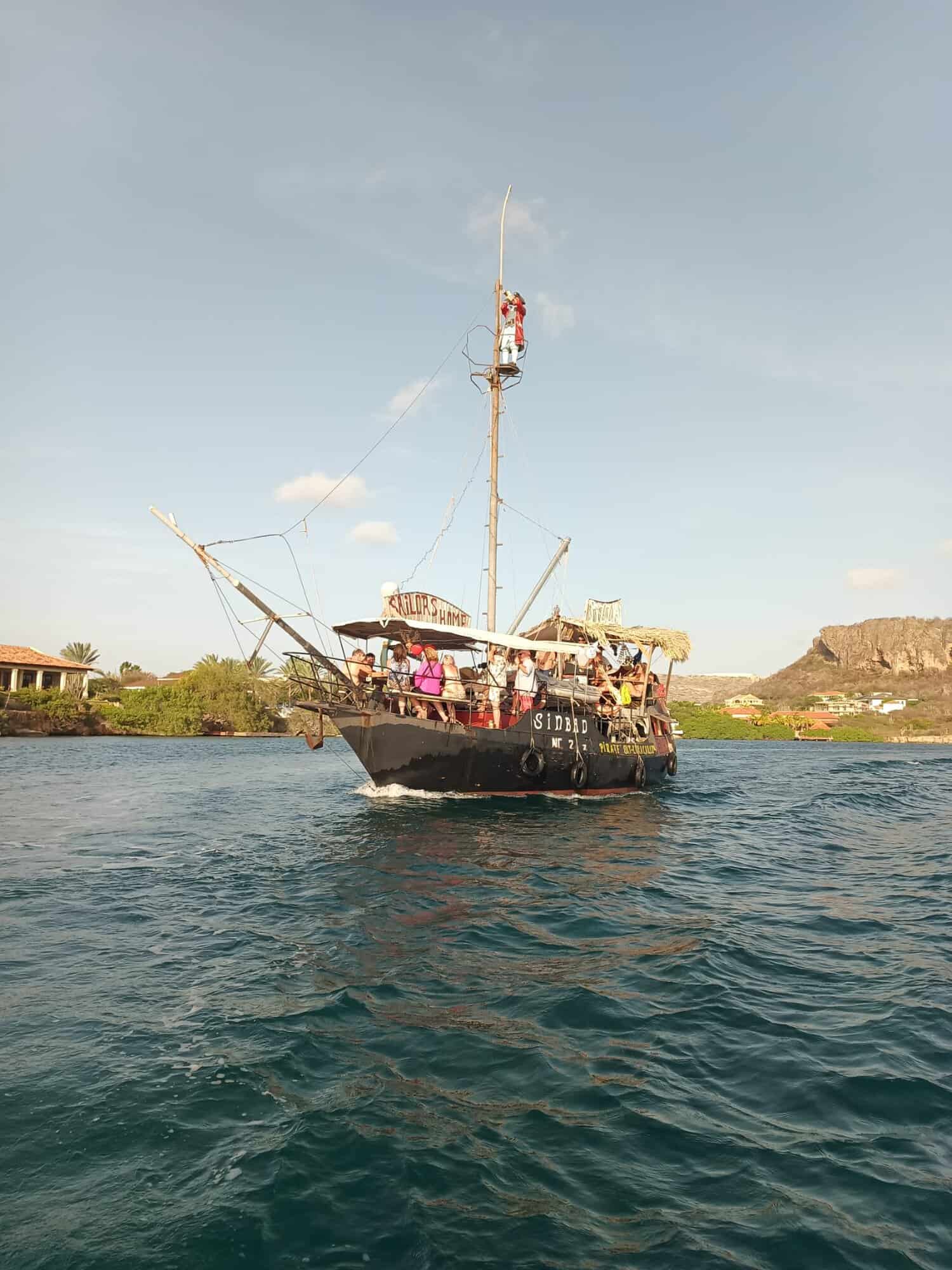 El barco negro de estilo pirata Simba ofrece el crucero Aventura pirata, navegando por aguas azules junto a una costa verde. Un hombre sube al mástil mientras la gente a bordo disfruta de las vistas de casas y una colina rocosa bajo un cielo despejado.