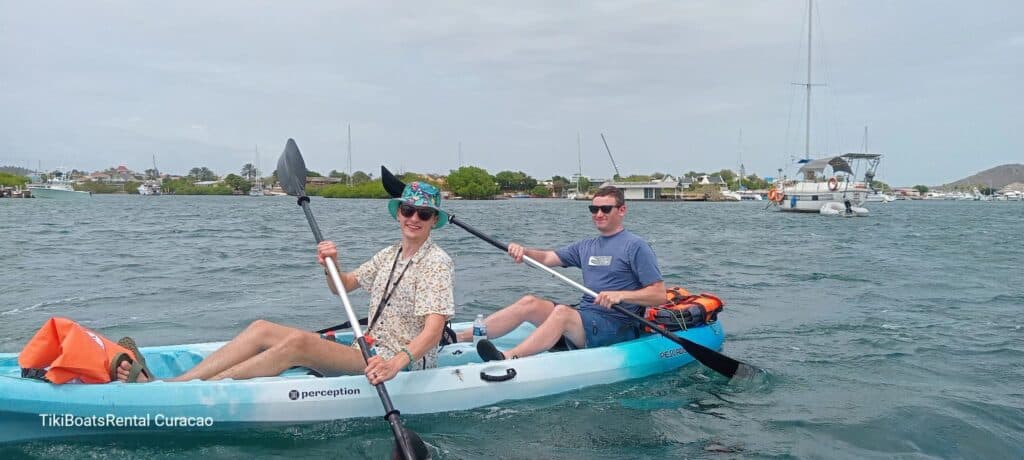 Two people in sunglasses and casual wear paddle the blue and white Tiki Kayak: Discover the Spanish Waters, with "TikiBoatRental Curacao" on its side. Boats and greenery surround them under a cloudy sky for a serene adventure.