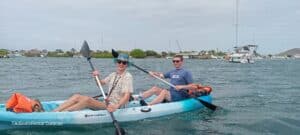 Two people in sunglasses and casual wear paddle the blue and white Tiki Kayak: Discover the Spanish Waters, with "TikiBoatRental Curacao" on its side. Boats and greenery surround them under a cloudy sky for a serene adventure.