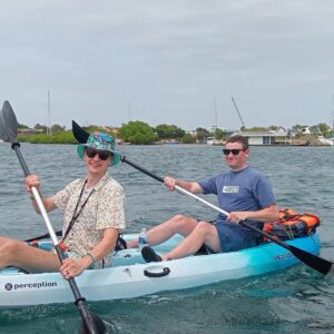 Dos hombres reman en un Kayak Tiki azul claro: Descubre las Aguas Españolas en aguas tranquilas, con barcas y una costa lejana. Ambos llevan gafas de sol; el de delante lleva un colorido sombrero Tiki y una camisa estampada, el de detrás va vestido de azul.