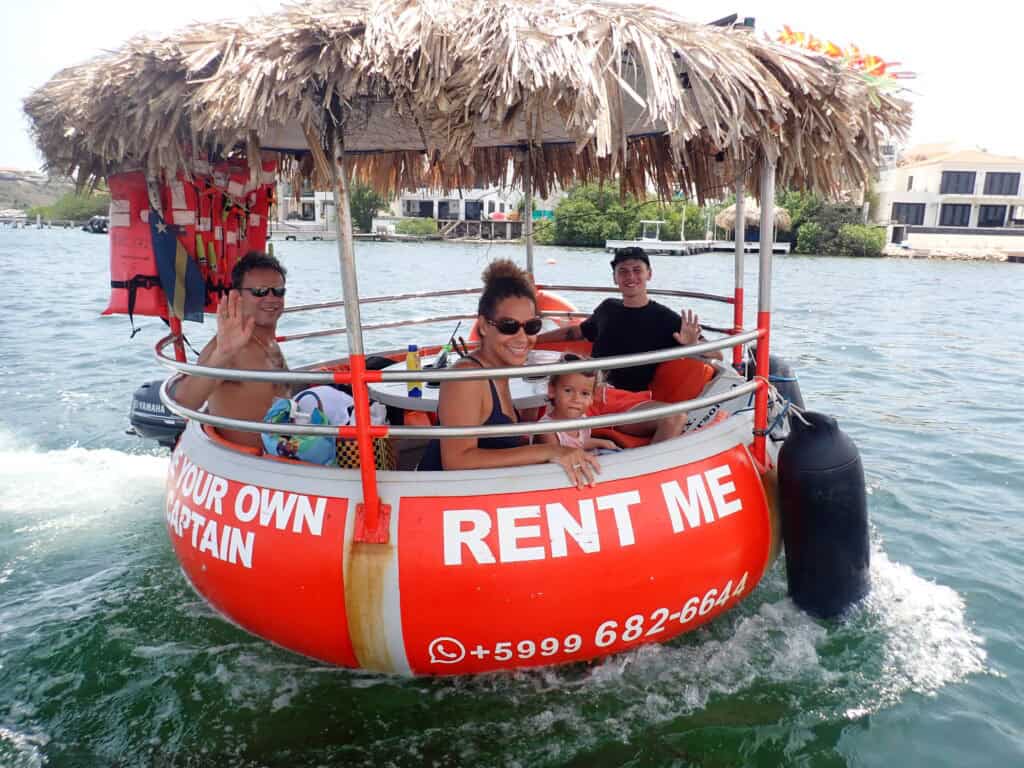Four people, including a child, smile and wave on a round red Tiki BBQ Boat Small: Be Your Own Captain with a thatched roof and “RENT ME” sign, cruising the water with houses visible in the background.