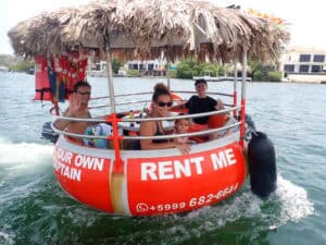 Four people, including a child, smile and wave on a round red Tiki BBQ Boat Small: Be Your Own Captain with a thatched roof and “RENT ME” sign, cruising the water with houses visible in the background.