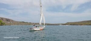 A small white Tiki Sailboat: The Ultimate Curacao Sailing Experience sails with four people on calm blue water near a green, hilly Curacao shoreline. Other boats appear in the distance under a partly cloudy sky. Text reads TikiBoatsRental Curacao.