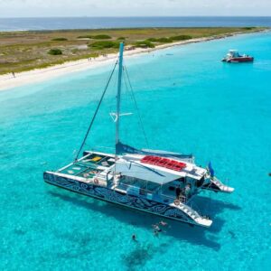 A colorful catamaran floats on clear turquoise water near a sandy beach with green vegetation, showcasing featured products as people swim, snorkel, and relax on boats under a sunny sky. Other boats are anchored nearby along the shoreline.